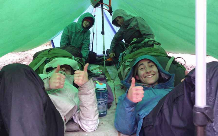 From under a tarp shelter, people smile and give the camera thumbs up.